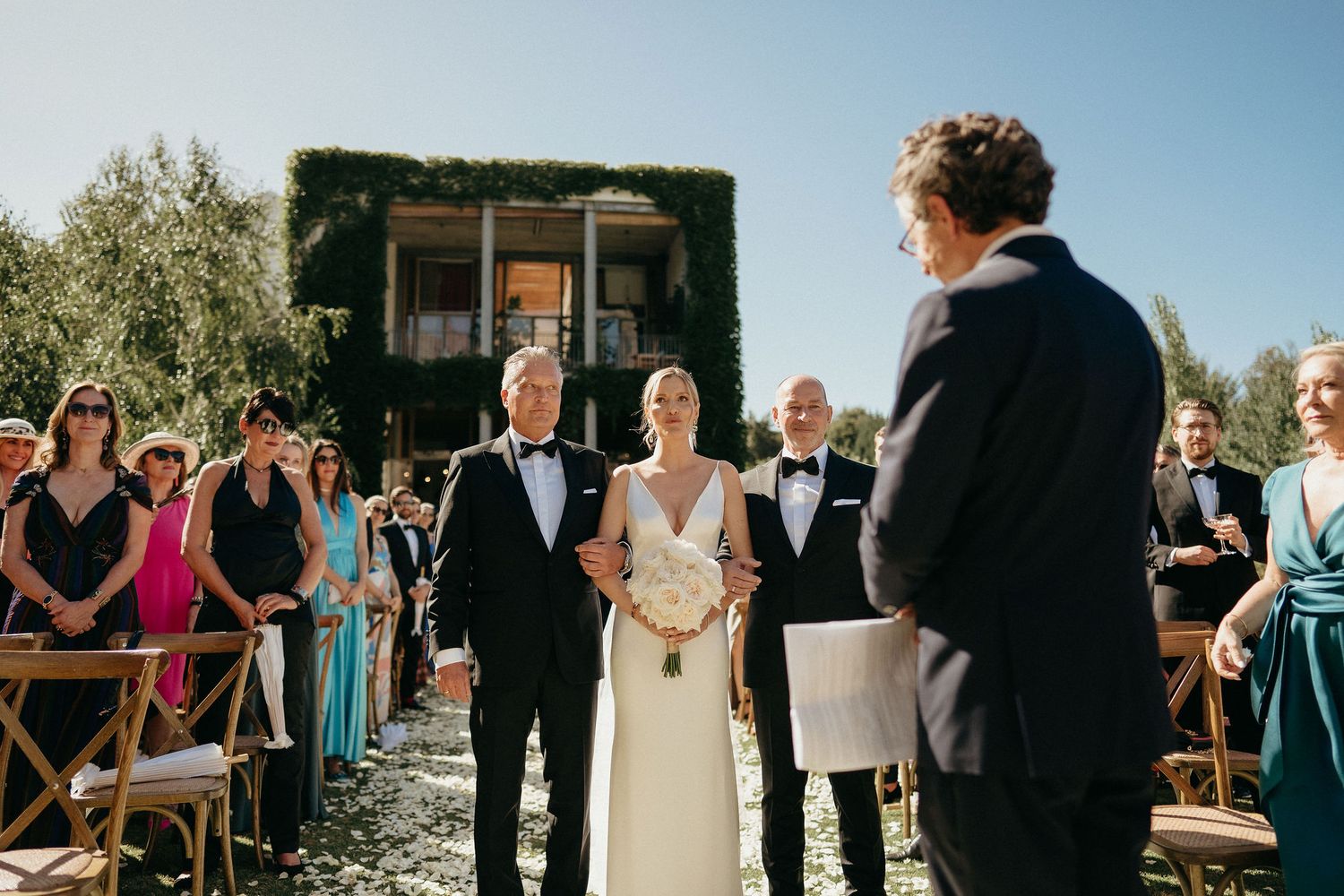 Wedding party gathered outdoors at scenic venue with guests seated in rows during sunny ceremony.