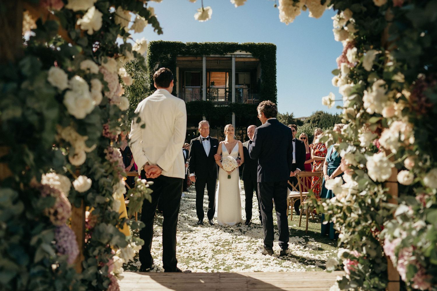 A wedding ceremony takes place under an elegant floral arch with white roses and natural sunlight streaming through.