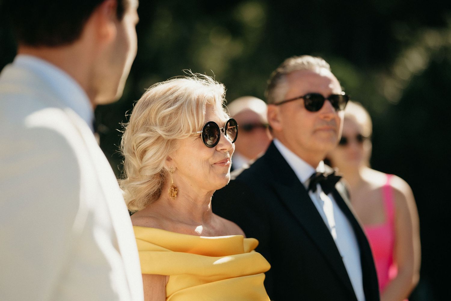 A group of formally dressed people stand together outdoors on a sunny day wearing sunglasses and formal attire.