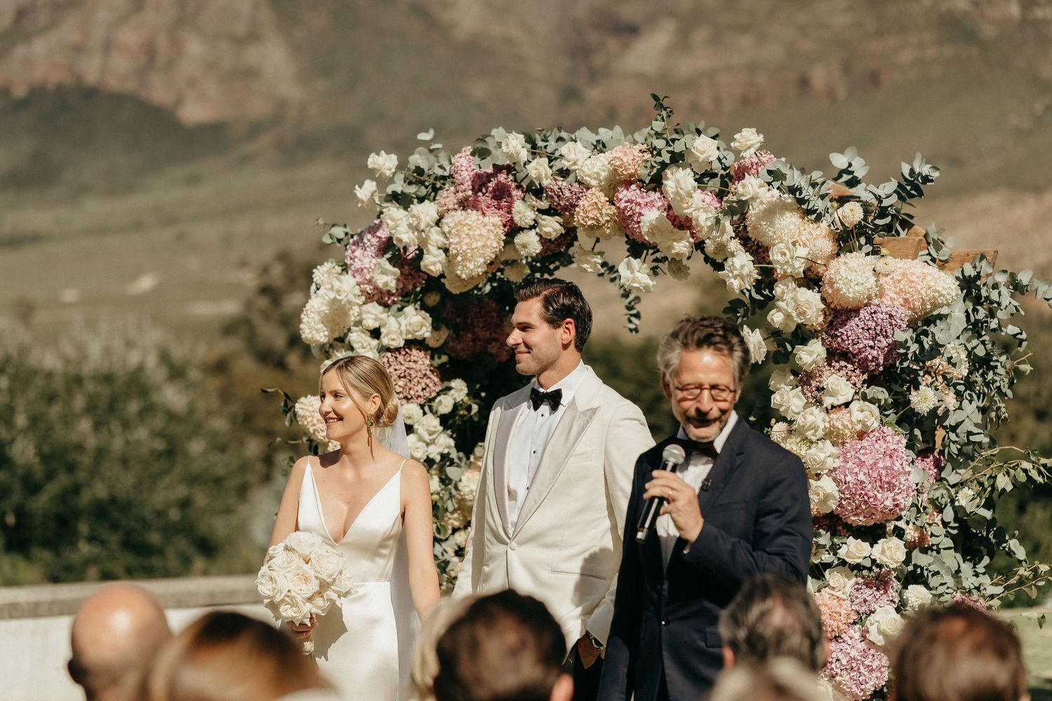 Wedding party stands in front of a floral arch with pink and white roses against a mountainous backdrop.
