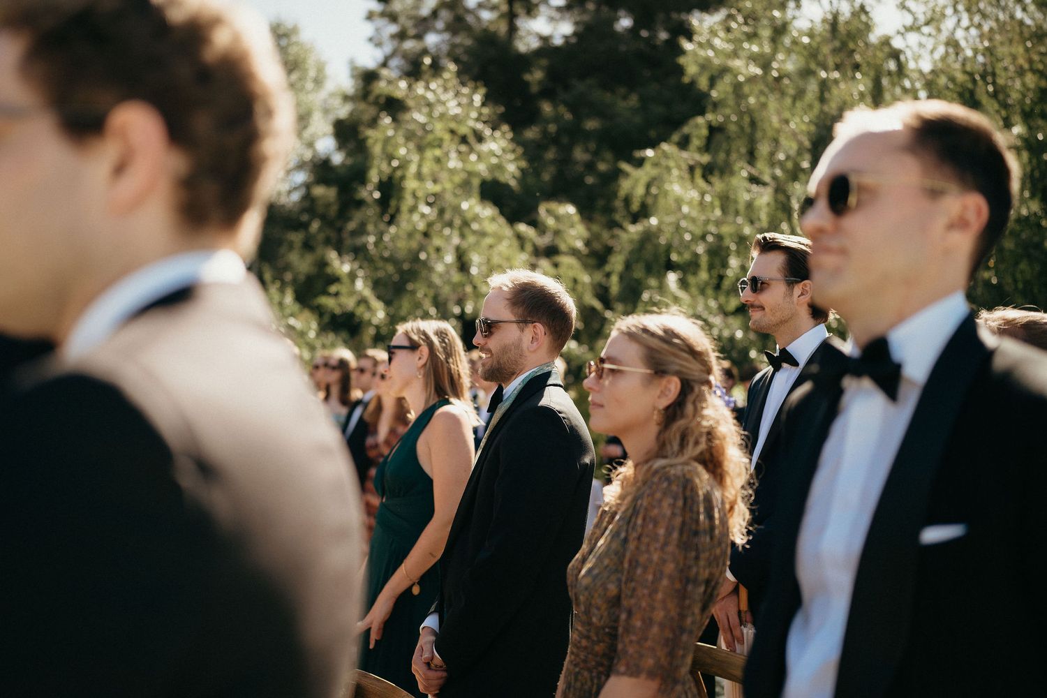 Wedding guests in formal attire stand outdoors watching a ceremony on a sunny day.