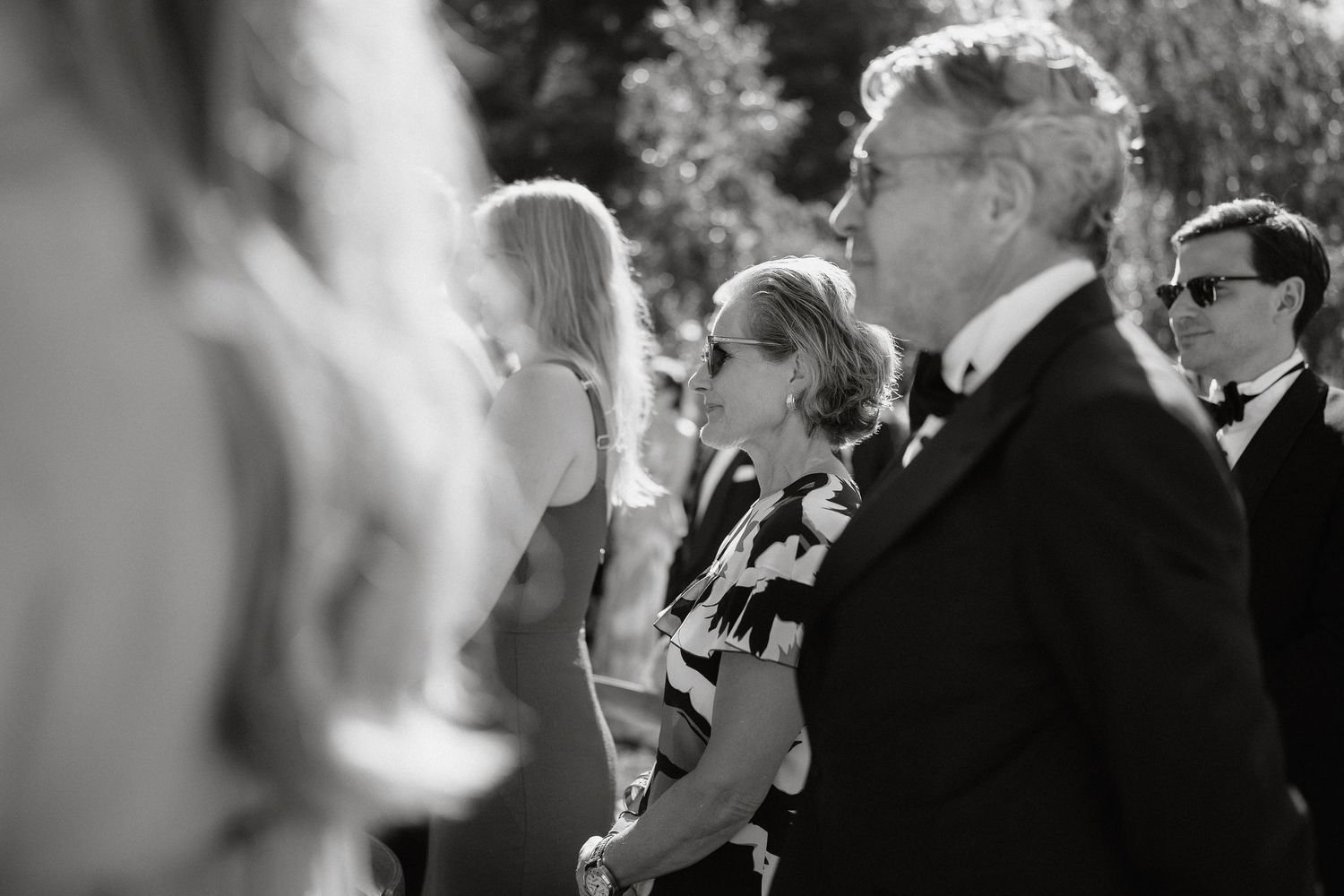 Black and white photo of wedding guests mingling and chatting at an outdoor reception celebration.