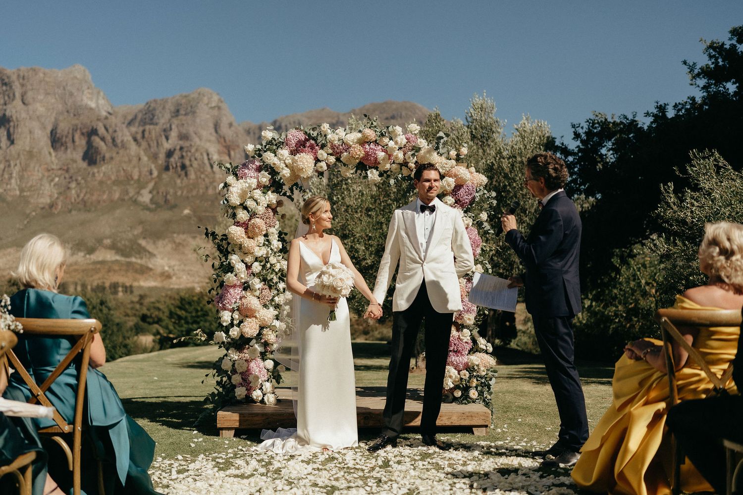 Desert wedding ceremony setup with rustic wooden chairs and pink floral arrangements against dramatic mountain backdrop.