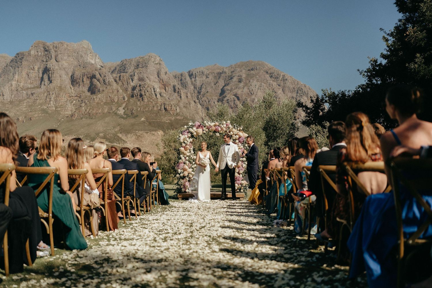 Outdoor desert wedding ceremony with rocky mountain backdrop and white aisle runner leading to floral arch.