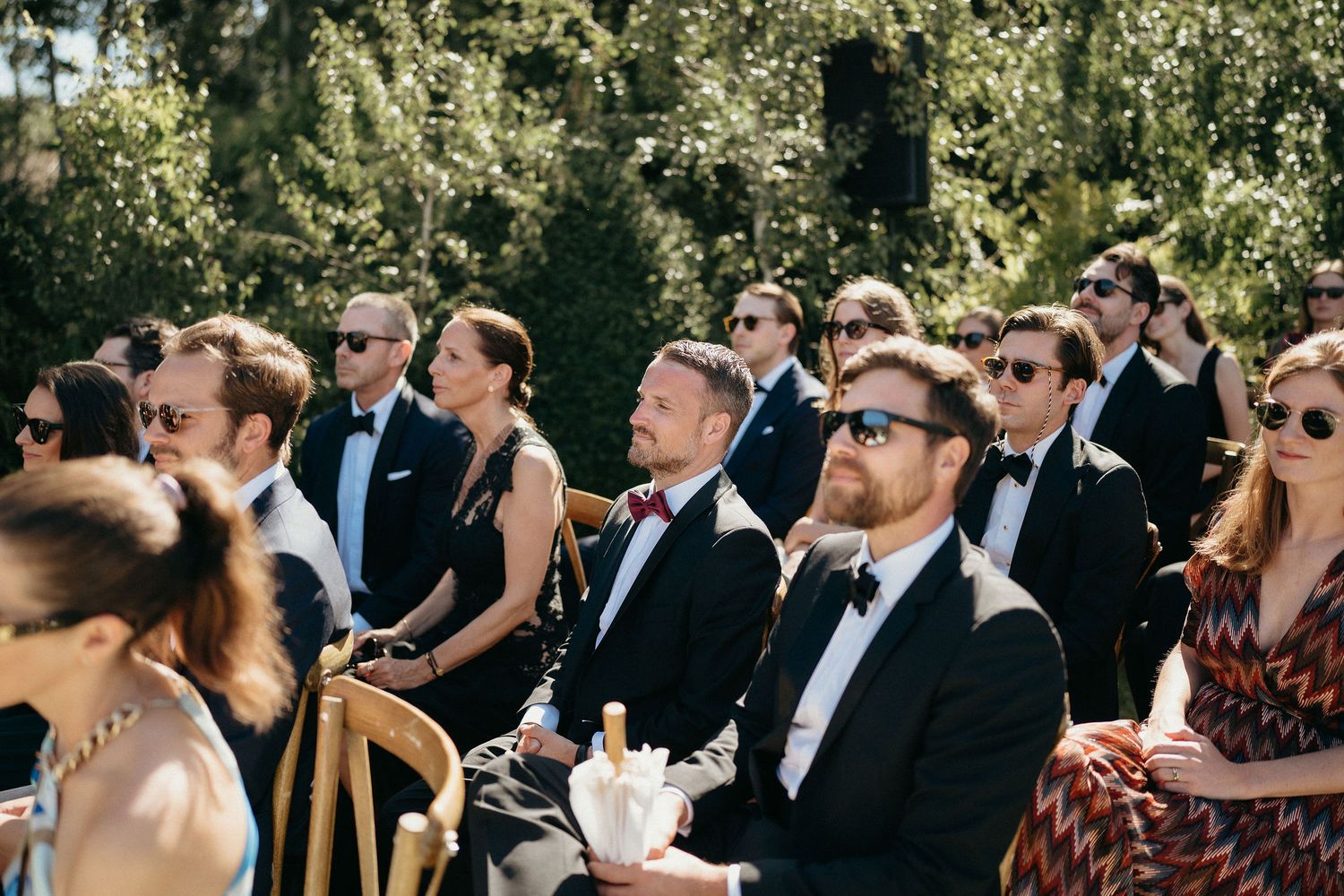 Wedding guests in formal attire seated outdoors during a sunny garden ceremony.