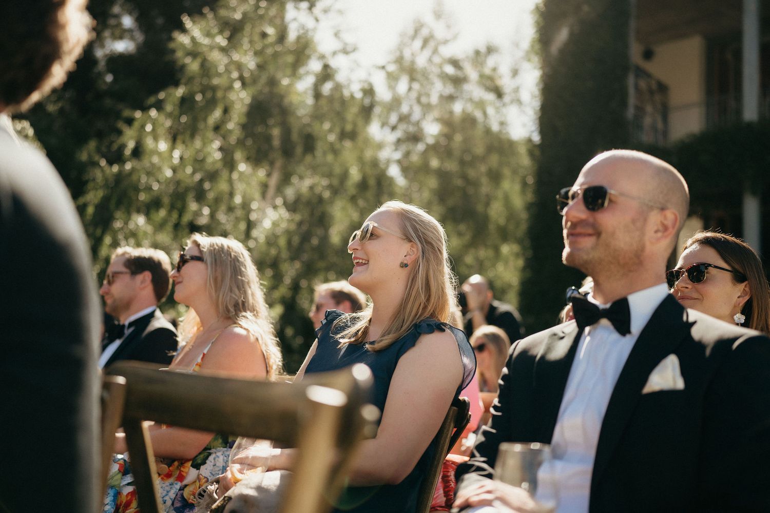 Wedding guests wearing sunglasses smile and look up at the sky during an outdoor celebration.