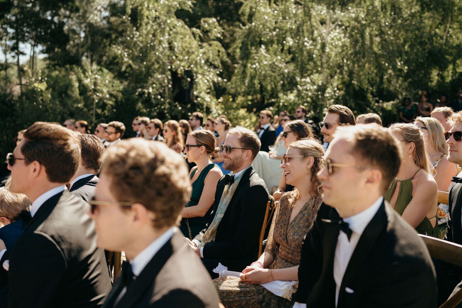A crowd of wedding guests in formal attire gathered outdoors on a sunny day during a celebration.
