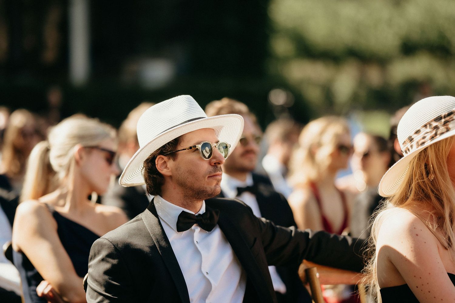 Wedding guests wearing cowboy hats and formal attire watch an outdoor ceremony on a sunny day.