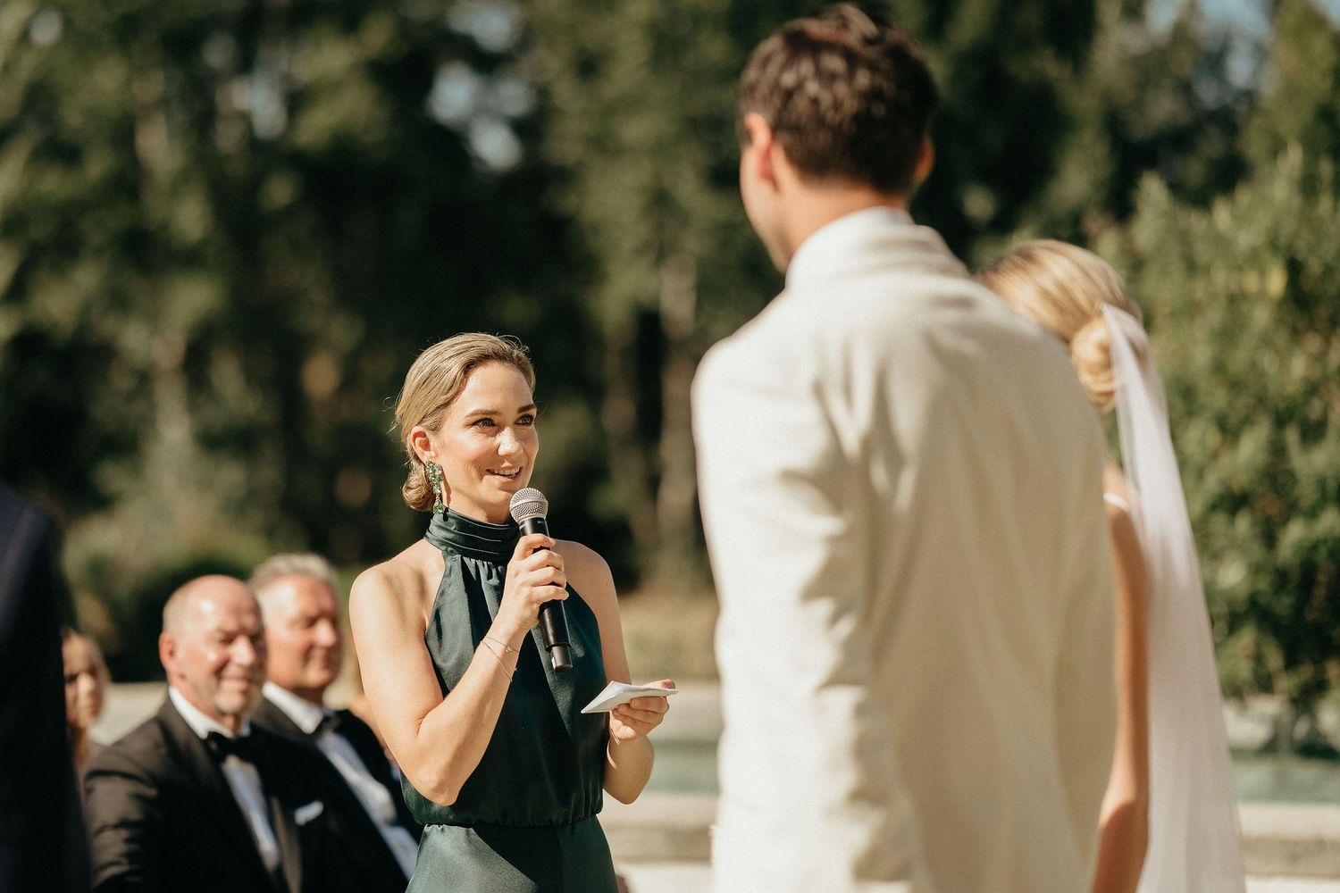 A joyful outdoor wedding ceremony with guests gathered under trees during a sunny summer day.