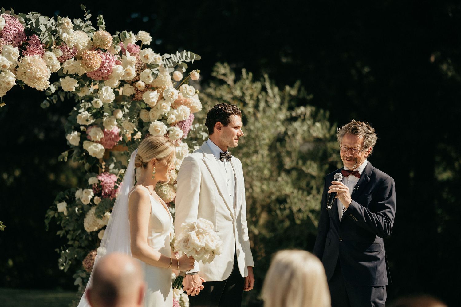 A romantic outdoor wedding ceremony with pink floral arrangements and guests gathered under sunny skies.