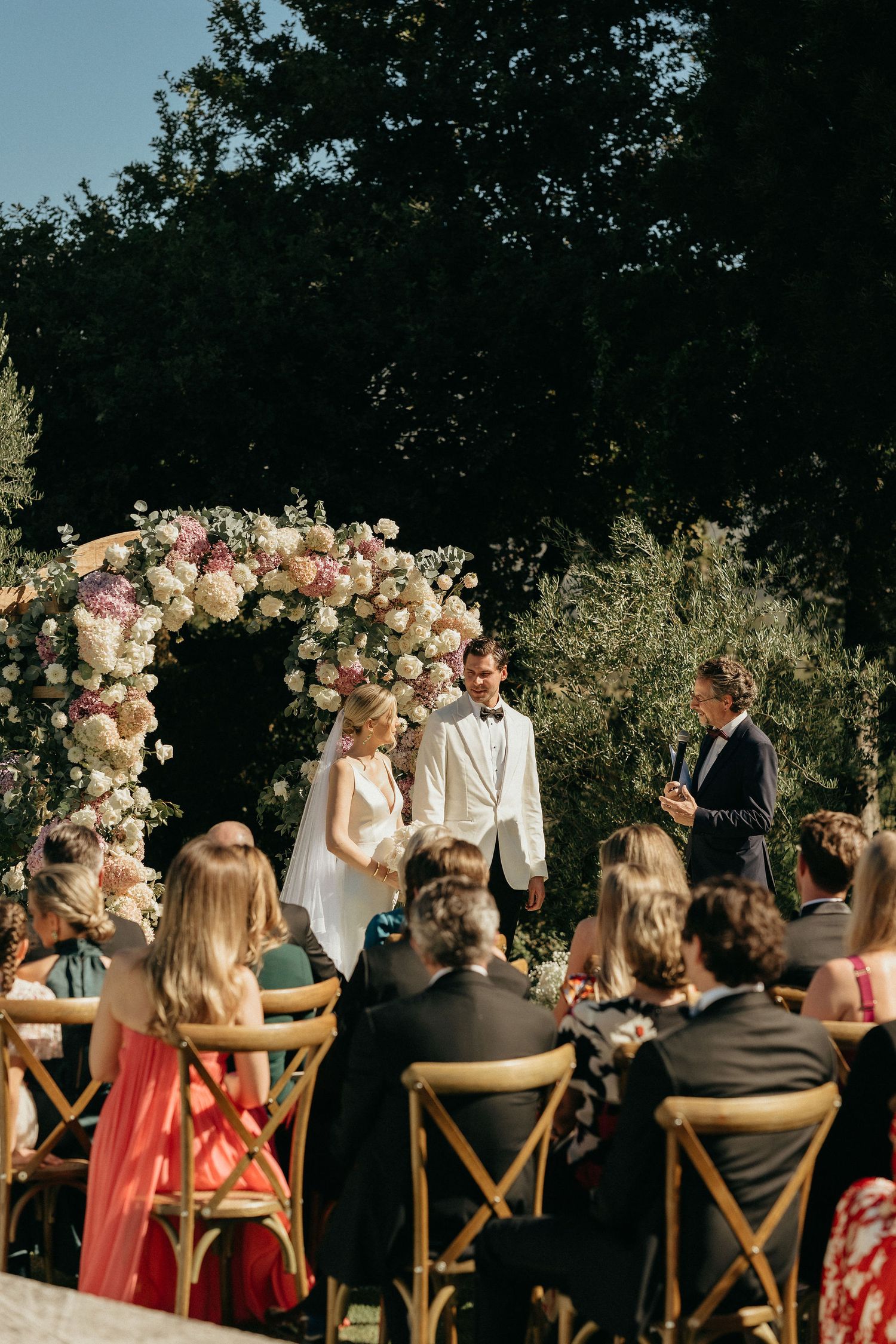 Wedding guests seated at rustic wooden chairs during an outdoor ceremony with floral arch decorations in natural setting.