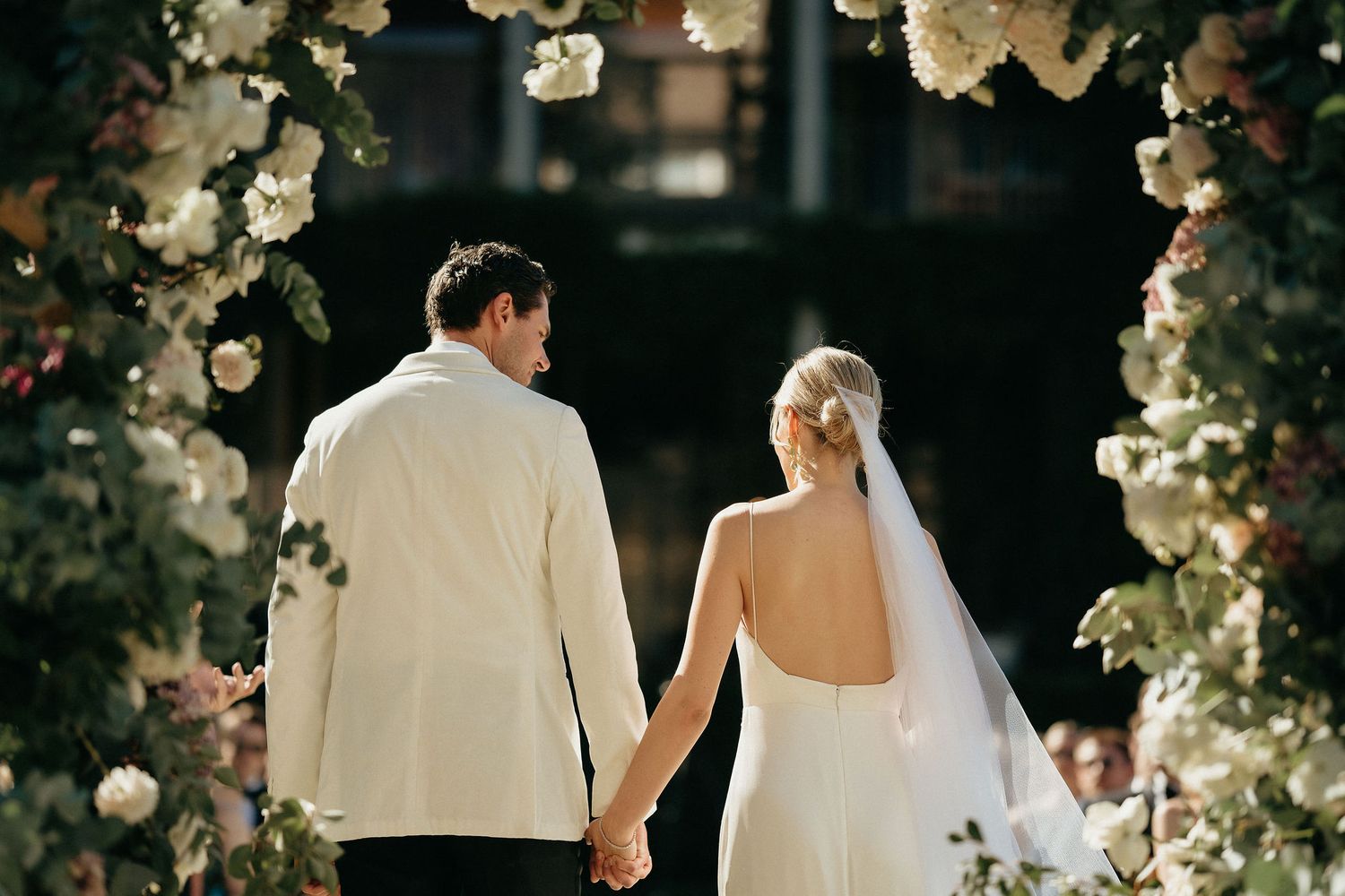 Couple in elegant white wedding attire standing beneath floral arch decorations during outdoor ceremony.