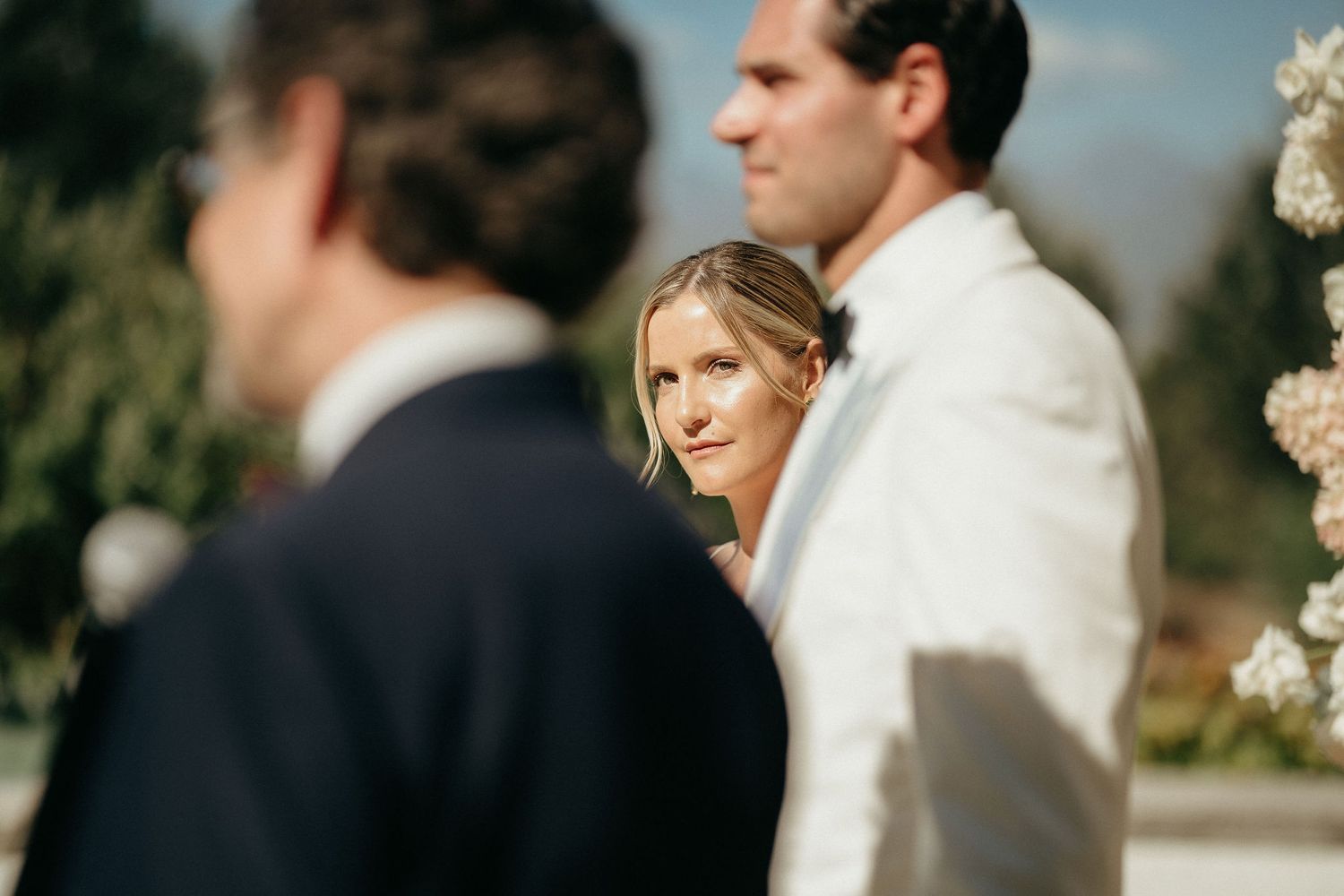 A couple in wedding attire stands close together during an outdoor ceremony with white floral decorations.