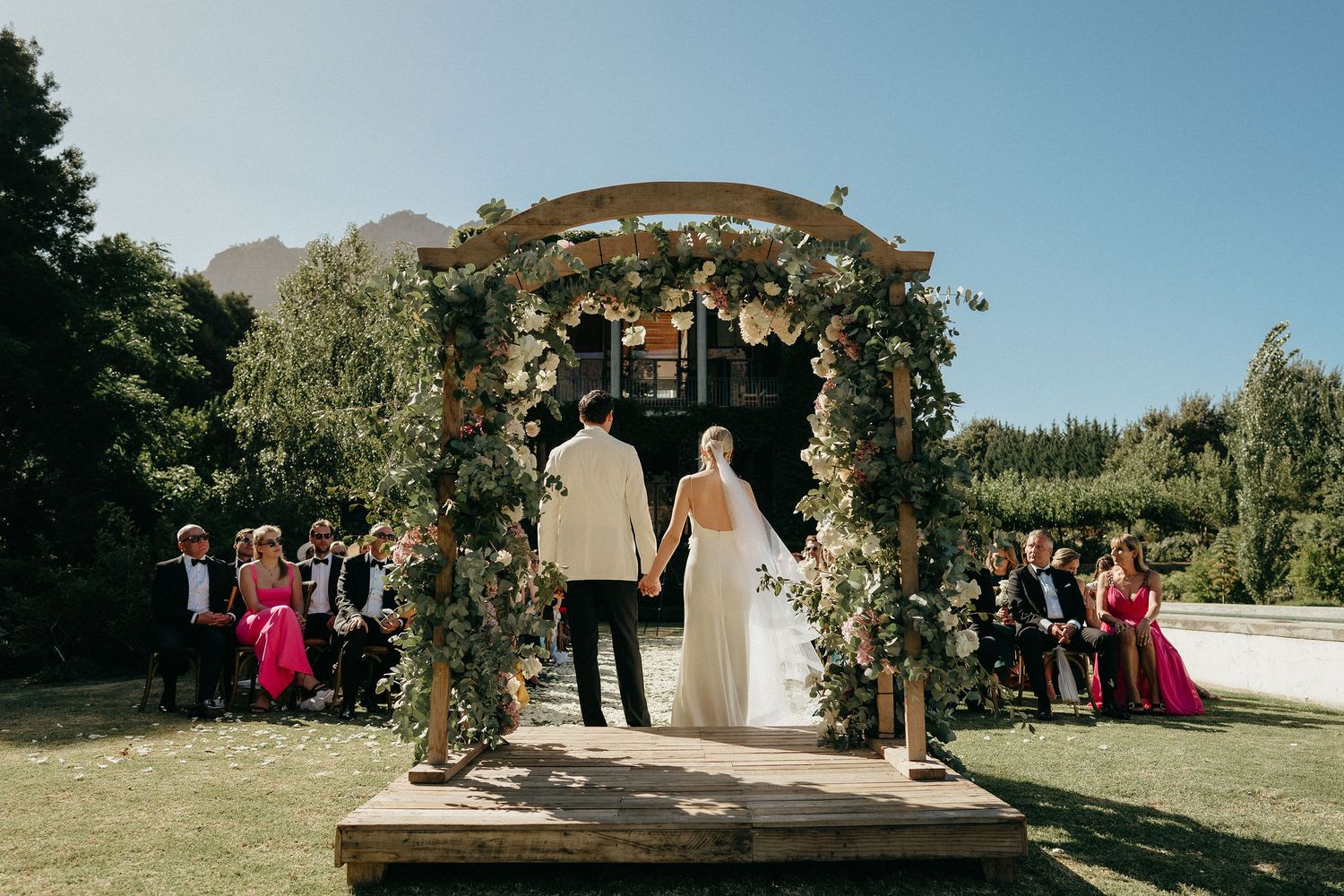 A rustic outdoor wedding ceremony setup with wooden arch and floral arrangements in a scenic garden setting.