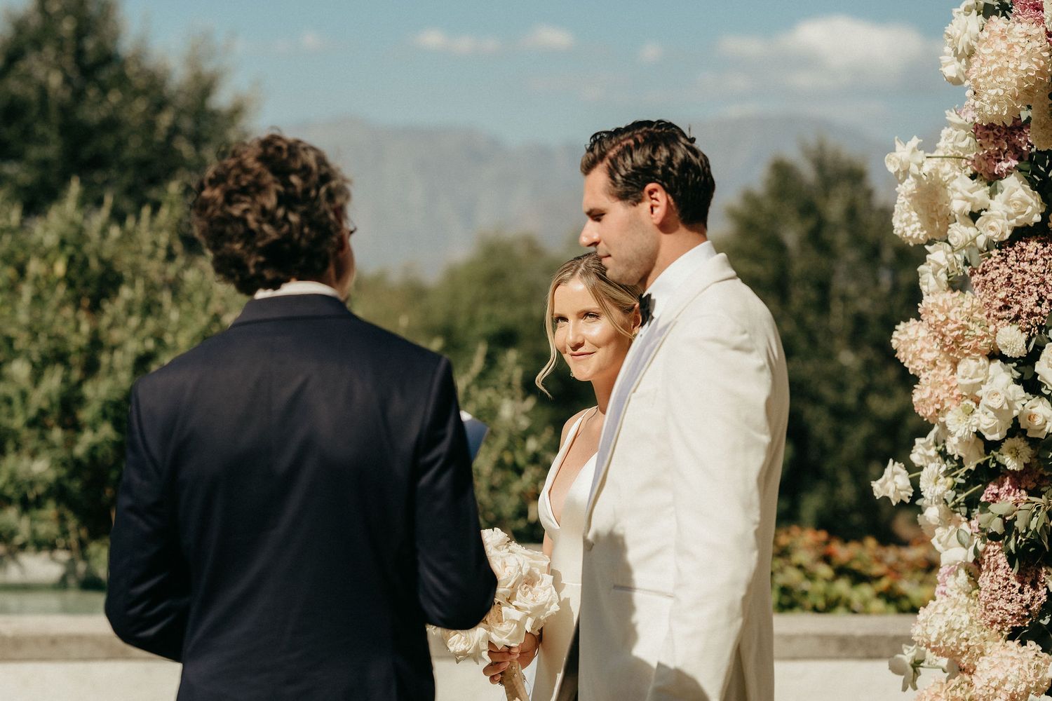 A wedding ceremony takes place outdoors with pink floral arrangements and mountain views in the background.