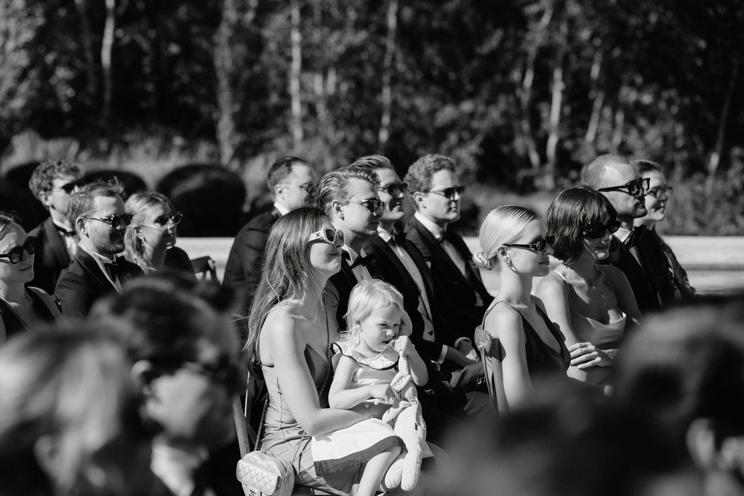 Black and white wedding ceremony photo showing guests seated outdoors in a natural setting during a summer celebration.