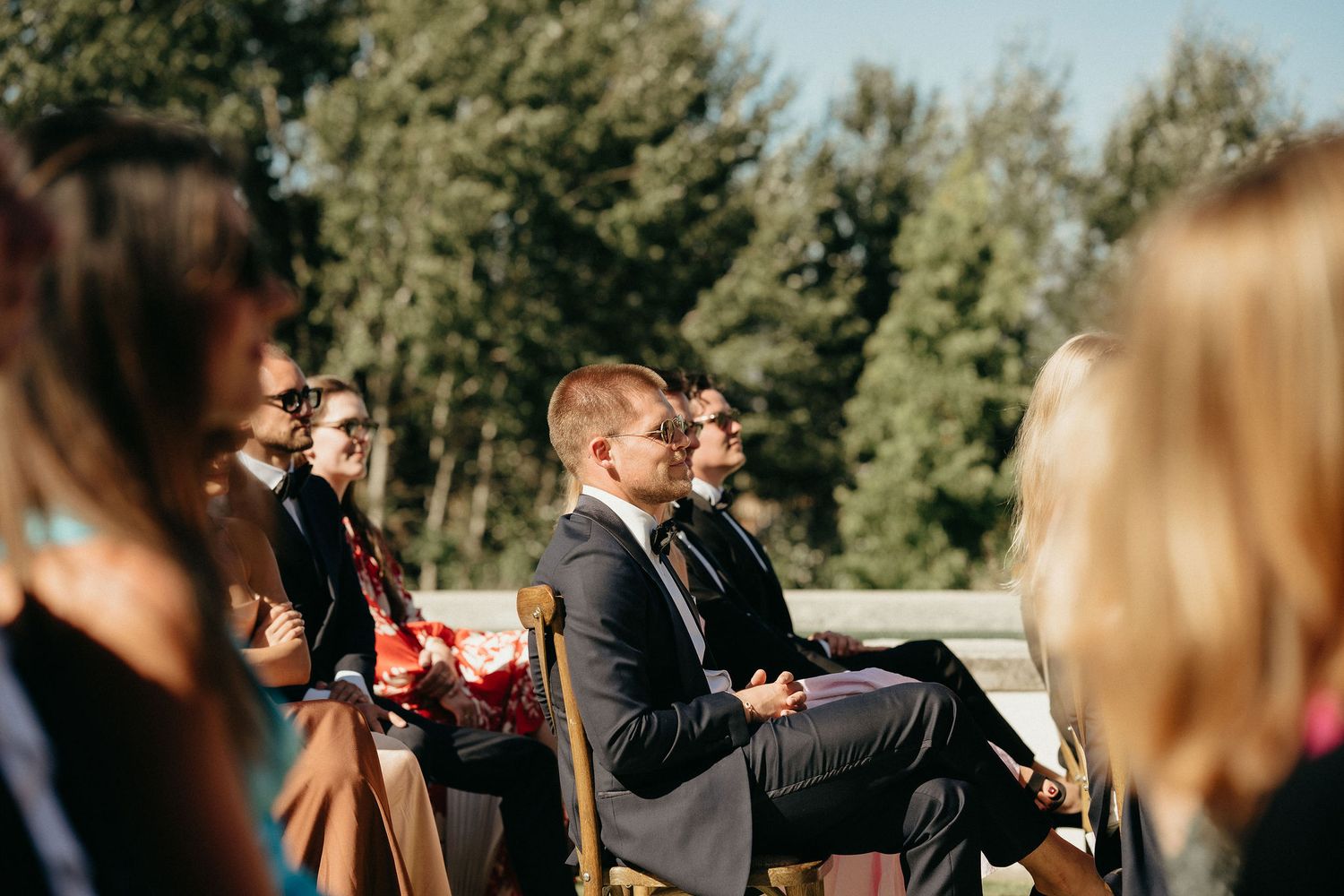 Wedding guests seated outdoors at a sunny ceremony with evergreen trees in the background.