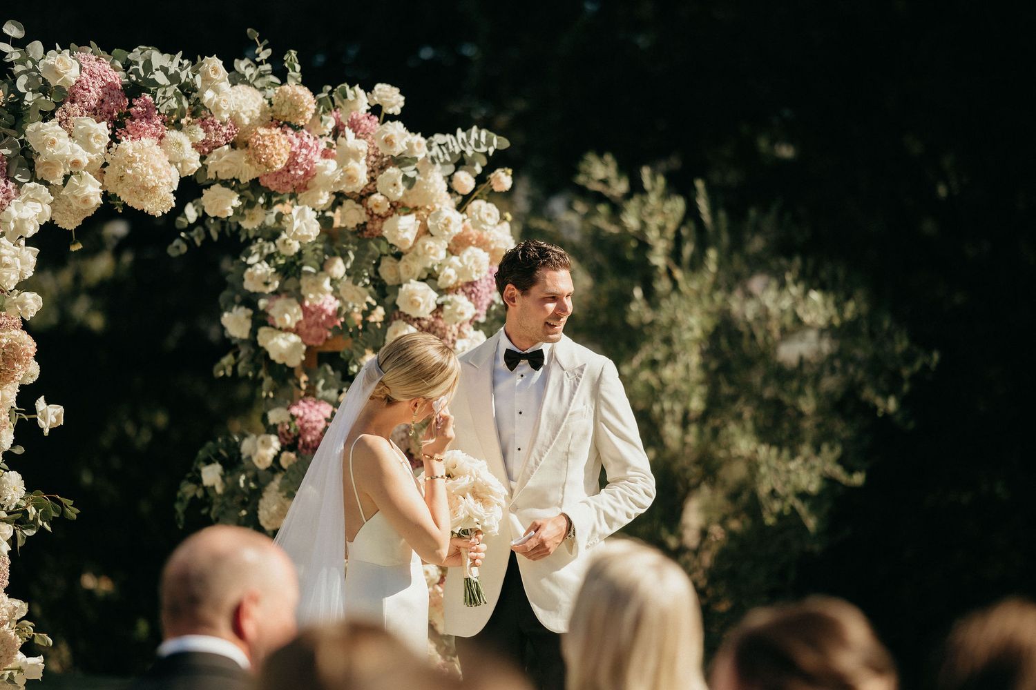 A romantic wedding ceremony under an elegant floral arch with pink and white roses in a garden setting.