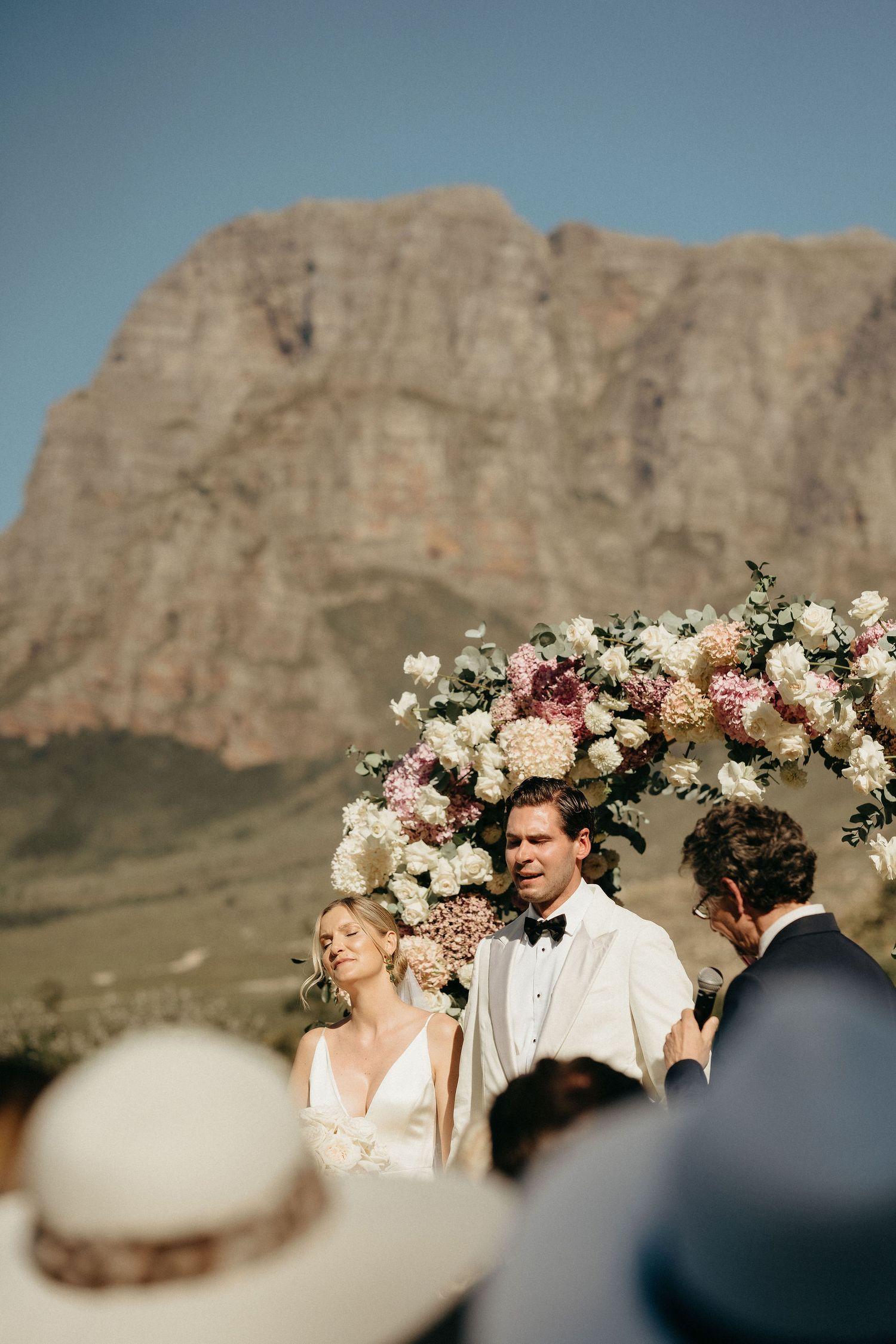 A scenic desert wedding ceremony takes place against dramatic red rock cliffs with floral arrangements in soft pink and white.