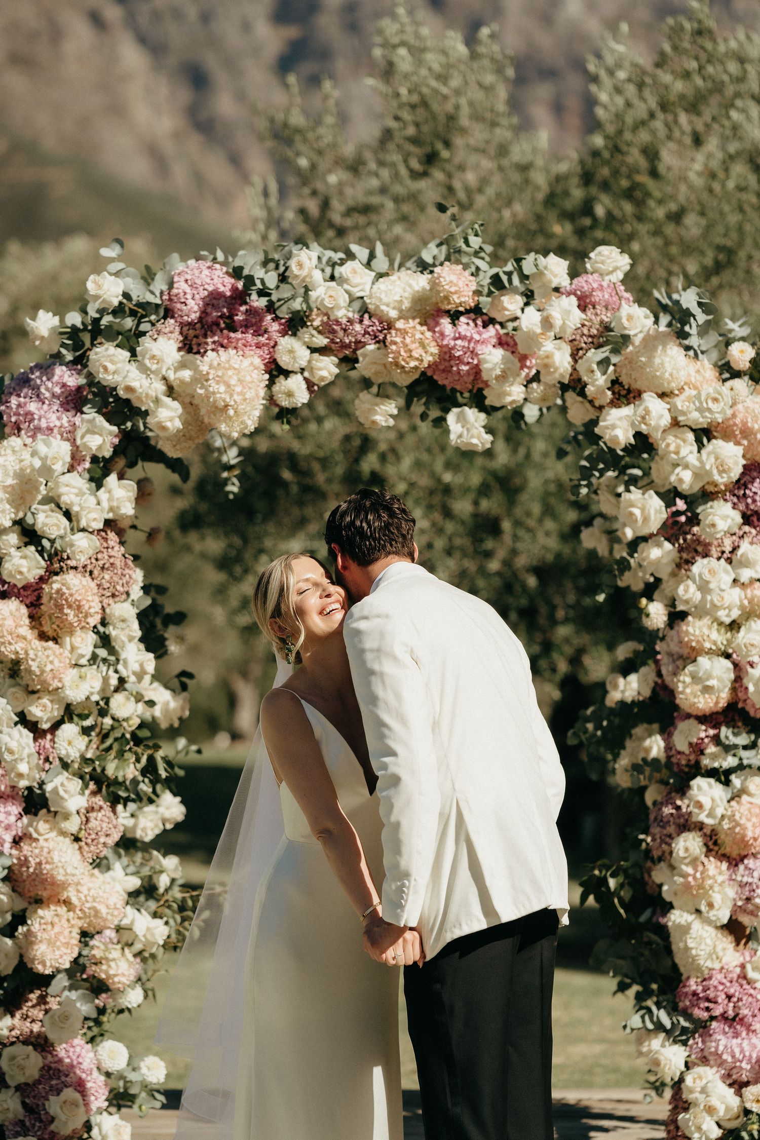 A romantic wedding ceremony moment under an elegant floral arch with pink and white roses in an outdoor setting.