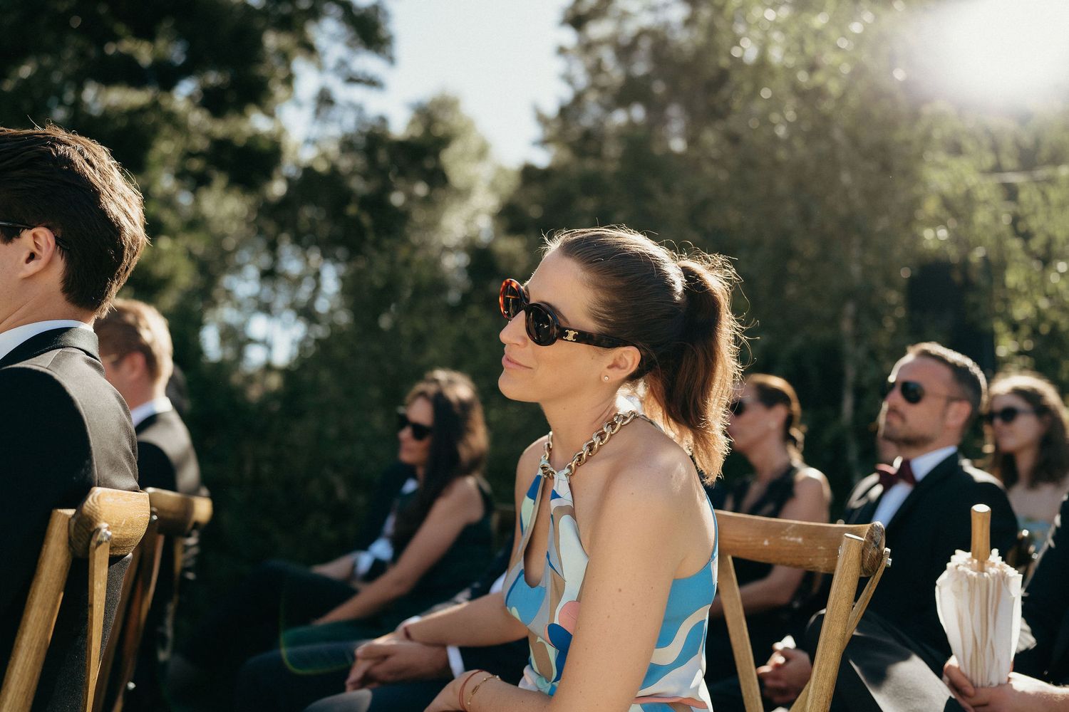 Crowd of wedding guests watching outdoor ceremony in sunlit garden setting.