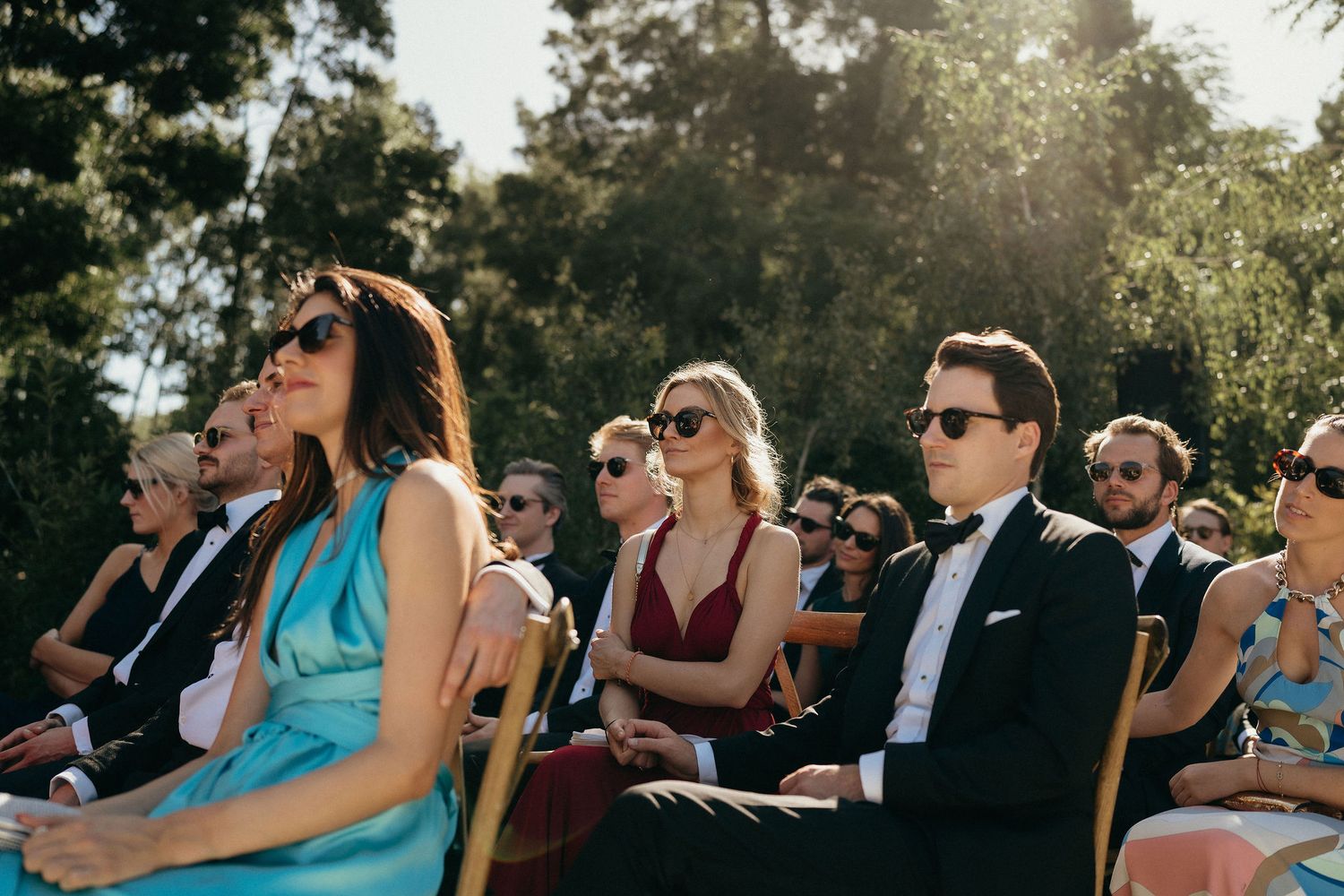 Wedding guests in turquoise dresses and formal attire sit in wooden chairs during an outdoor ceremony at sunset.