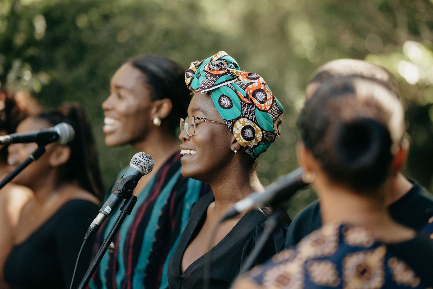 Wedding attendees seated outdoors wearing colorful headwraps and sunglasses.