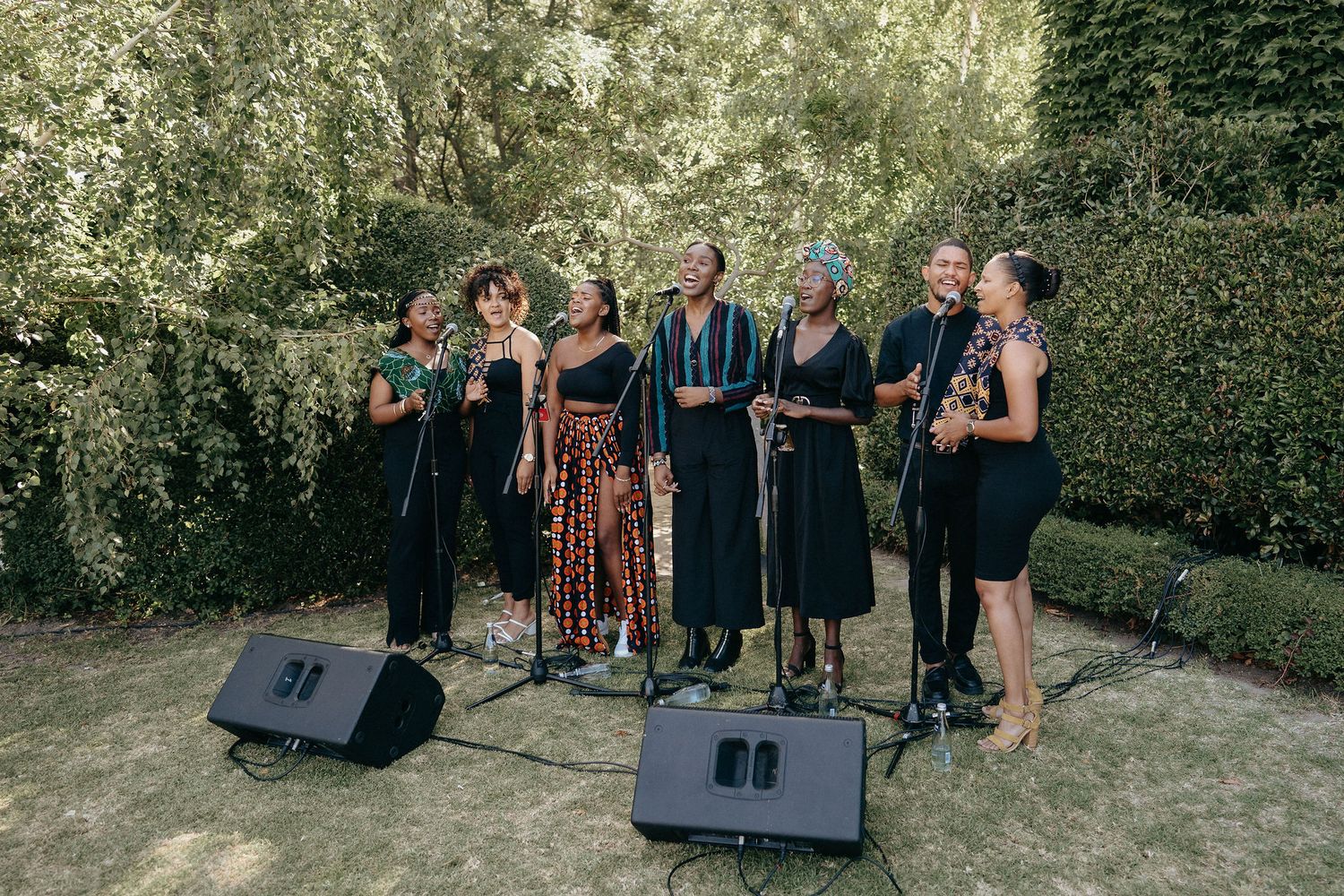 A cappella group performs outdoors with monitor speakers on a lawn against a green hedge backdrop.