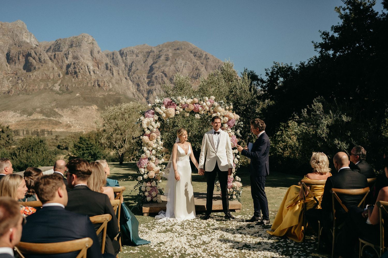 Scenic outdoor wedding ceremony with mountain backdrop and rustic wooden chairs arranged in rows.