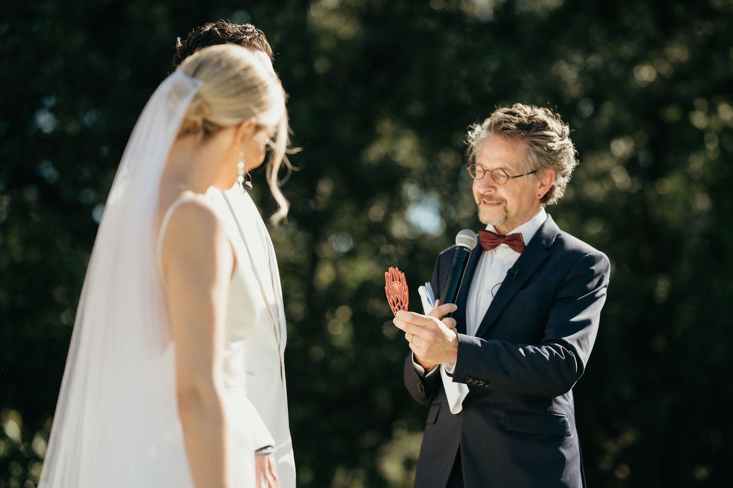 A couple shares an emotional moment during an outdoor wedding ceremony as the groom gestures while speaking.