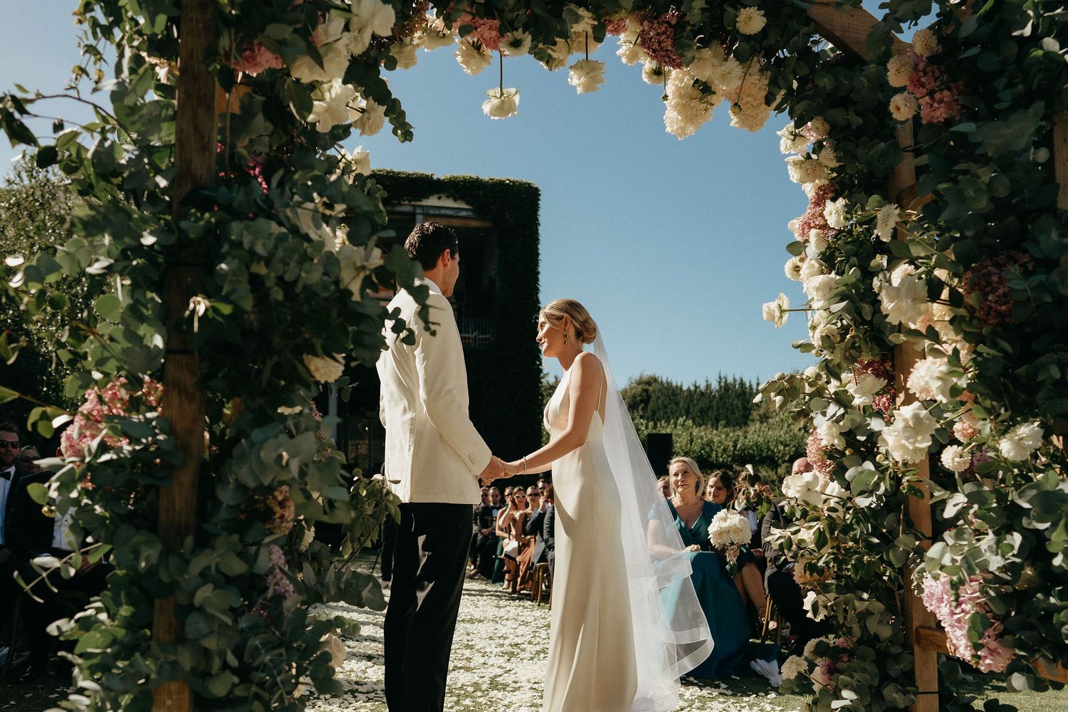 A couple shares a romantic moment under a floral arch during an outdoor wedding ceremony at sunset.