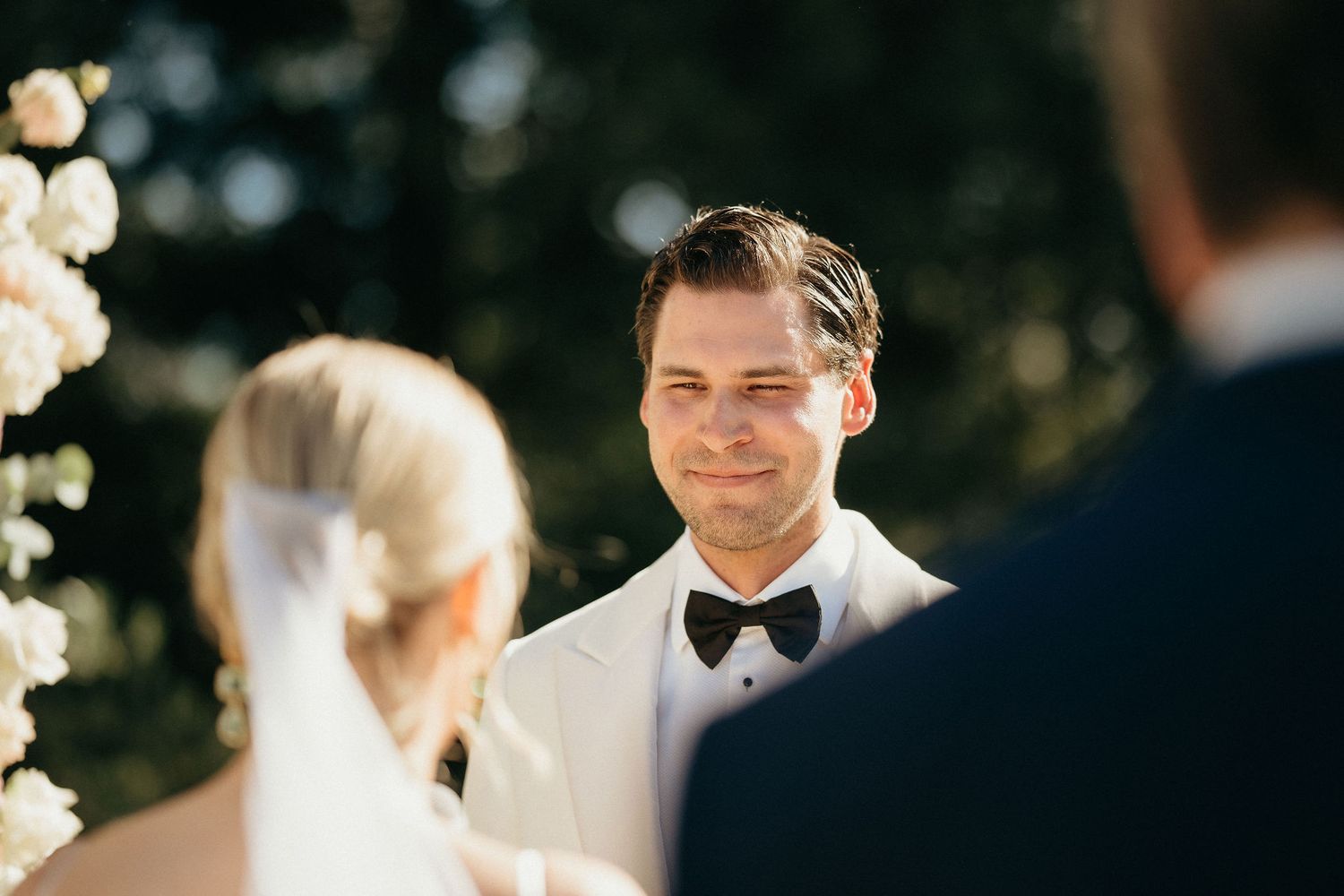 A couple exchanges rings during an outdoor wedding ceremony under bright sunlight.