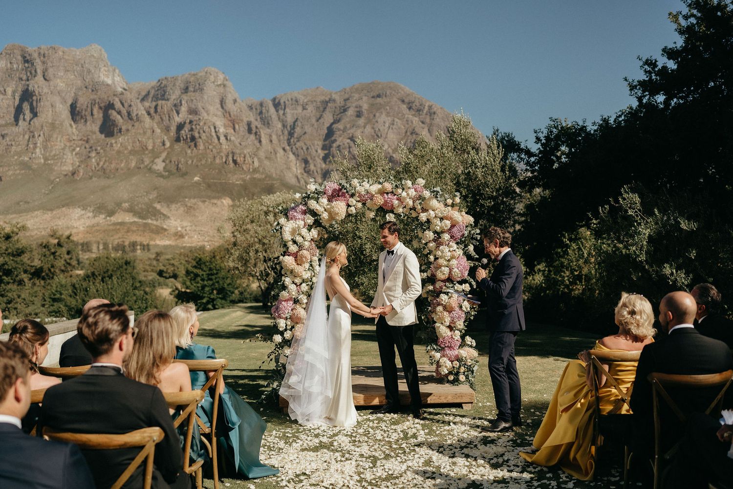 A scenic mountain wedding ceremony takes place outdoors by a serene lake with rocky cliffs in the background.