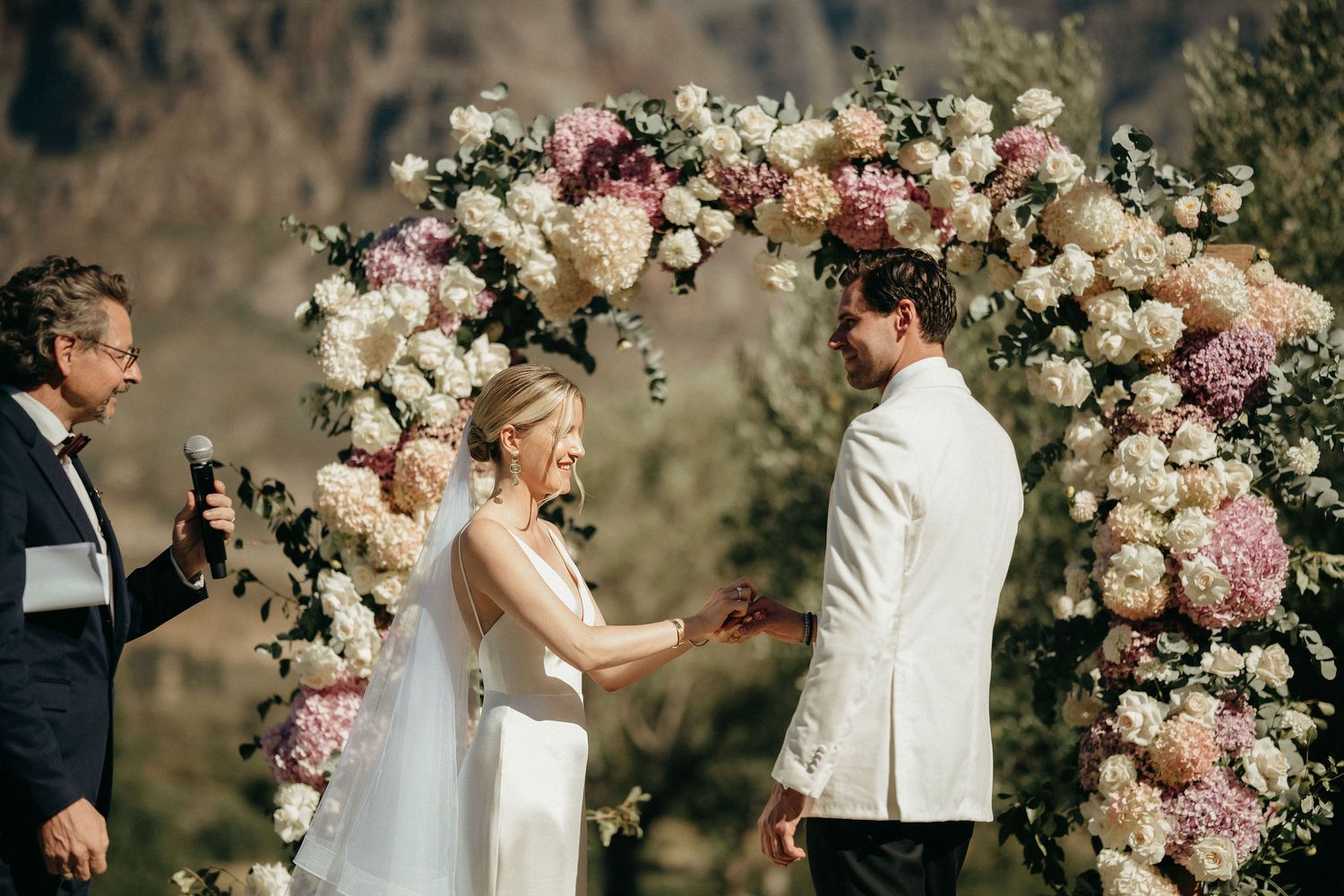 Wedding couple stands under a stunning floral arch with pink and white roses during an outdoor ceremony.
