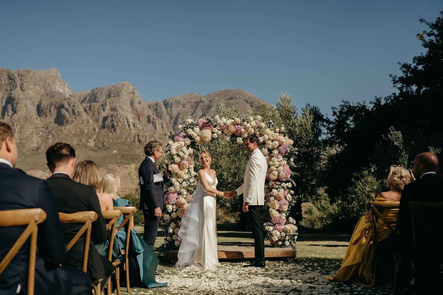 A wedding ceremony takes place in front of a stunning desert mountain backdrop with a floral arch arrangement.