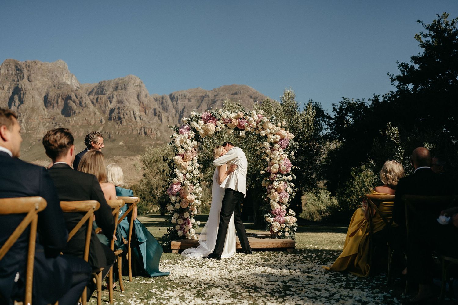 Outdoor wedding ceremony setup in front of majestic desert cliffs with wooden arch and white floral arrangements.