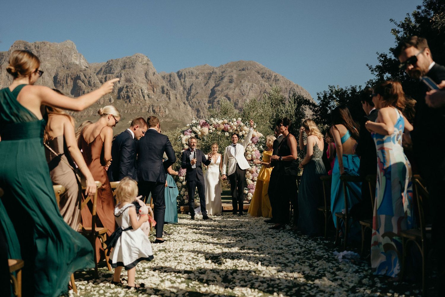 Wedding guests gather outdoors with dramatic mountain landscape in background.