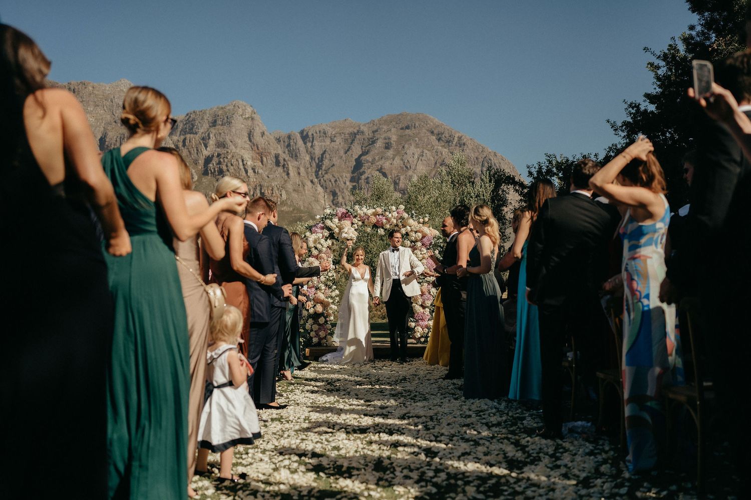 Wedding guests celebrate with flower petals against a scenic mountain backdrop in an outdoor ceremony.