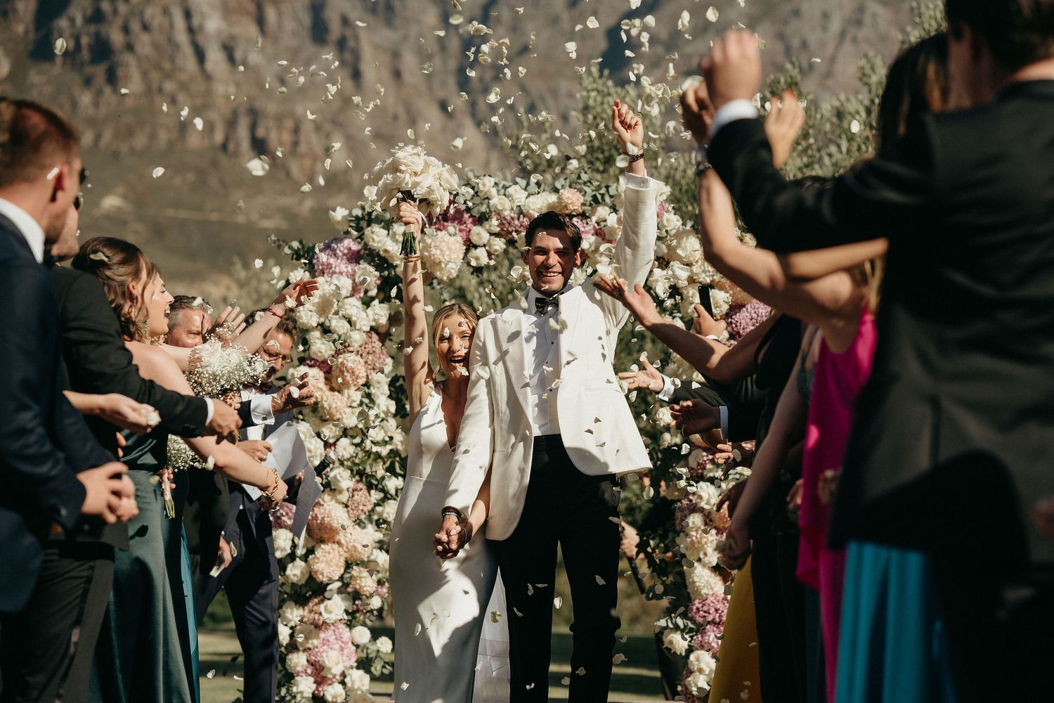 Guests toss pink flower petals during an outdoor wedding celebration with mountain views.