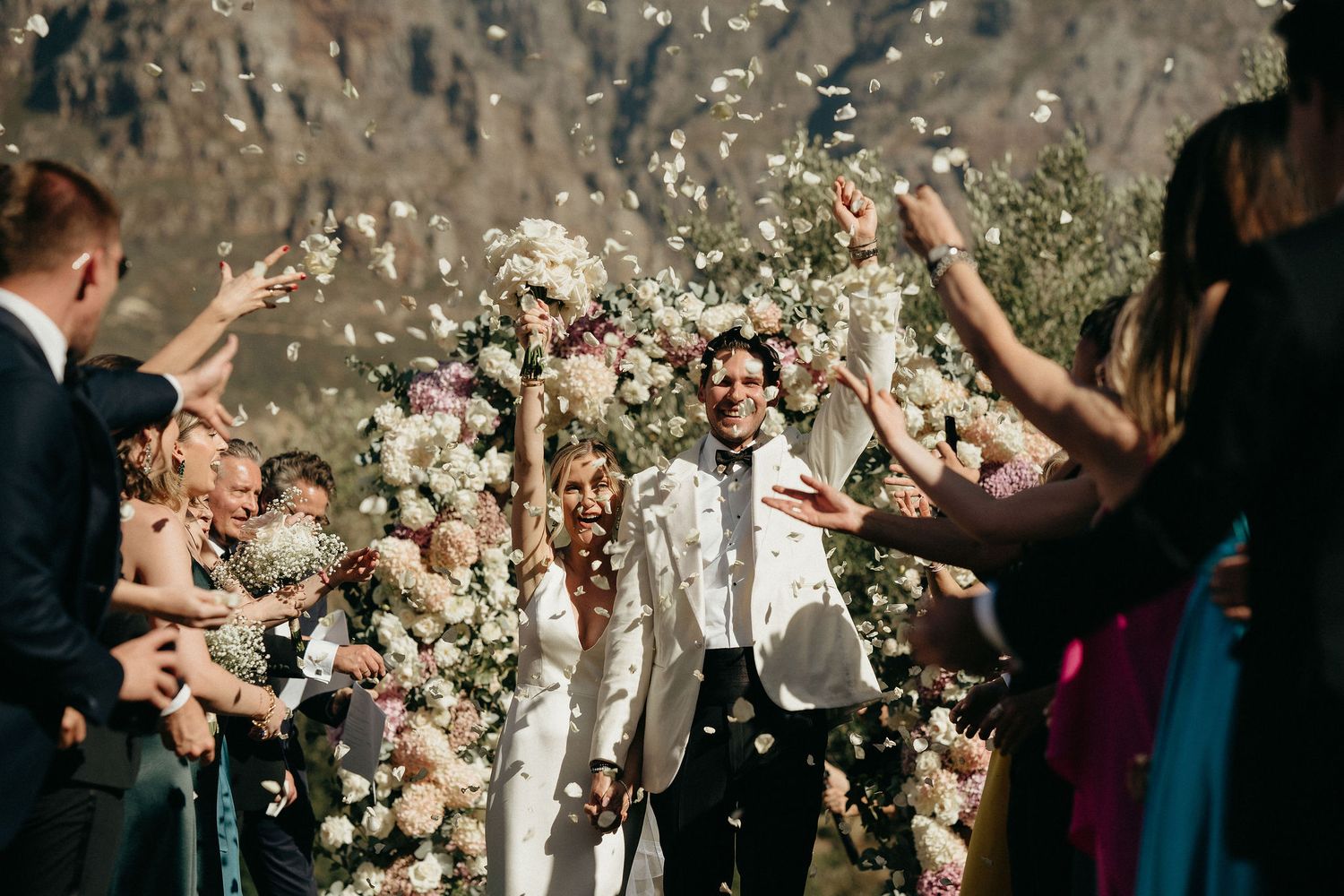 Wedding guests throw flower petals in celebration as newlyweds walk beneath a floral-decorated outdoor archway.