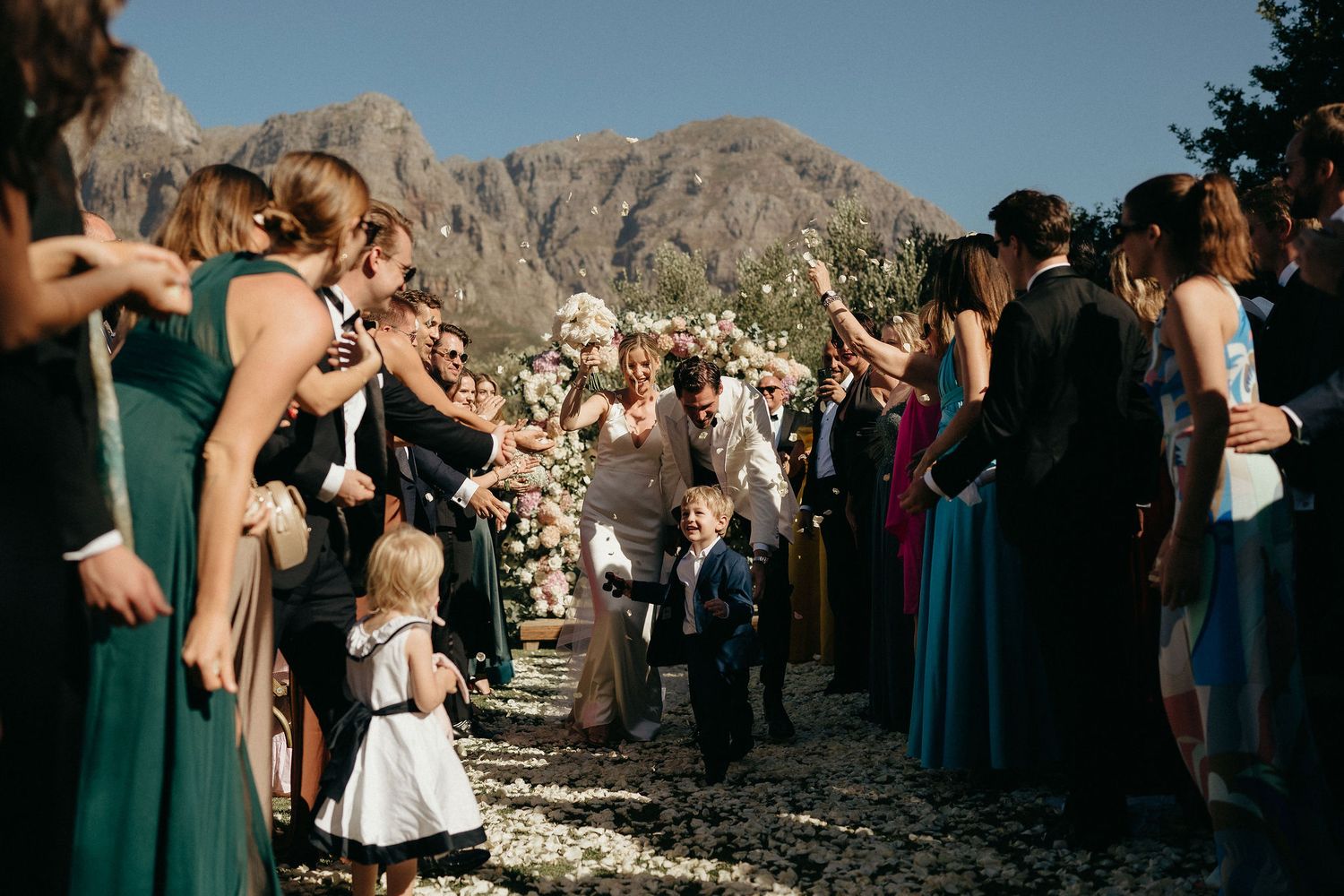Wedding guests gather in a scenic mountain setting with dramatic rocky cliffs as the backdrop during an outdoor ceremony.