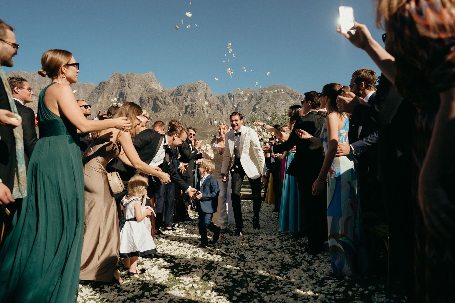 Wedding guests gather outdoors in green and beige dresses against a mountain backdrop during a celebration.