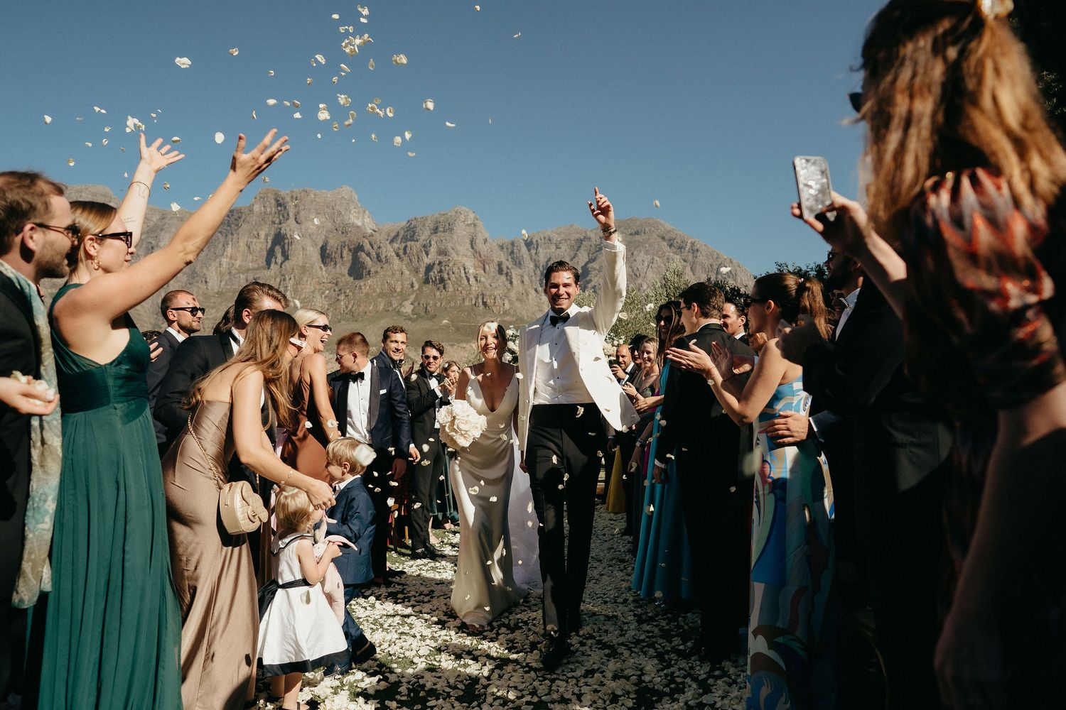 Wedding guests toss confetti over newlyweds during outdoor celebration with mountain backdrop and green-clad bridal party.