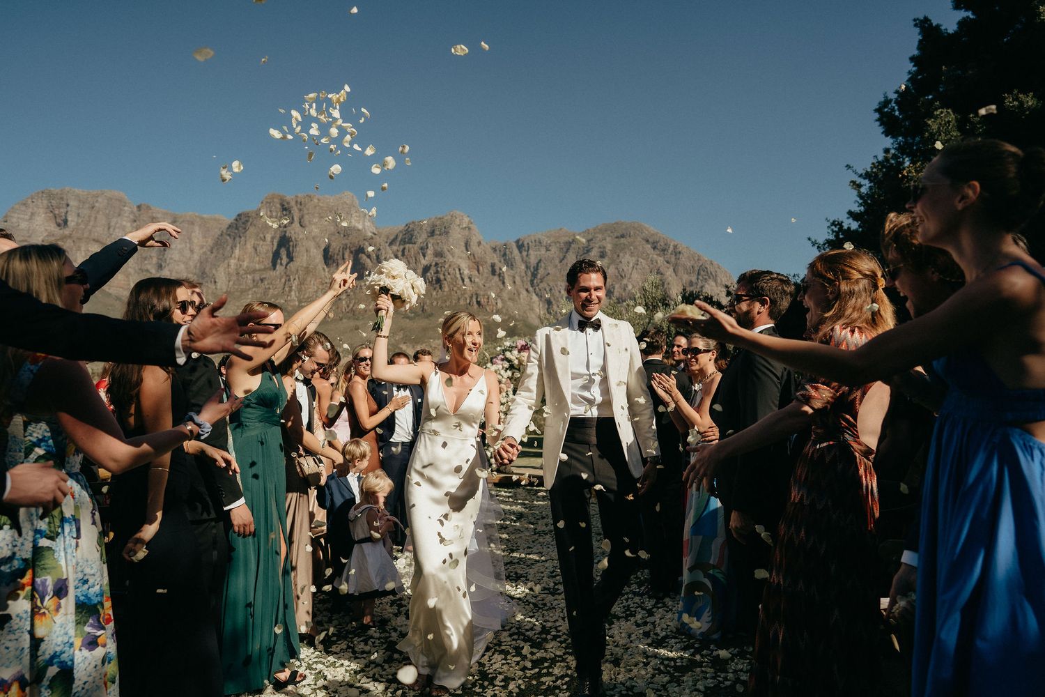 Wedding guests throw confetti at a joyous outdoor celebration with mountain peaks in the background.