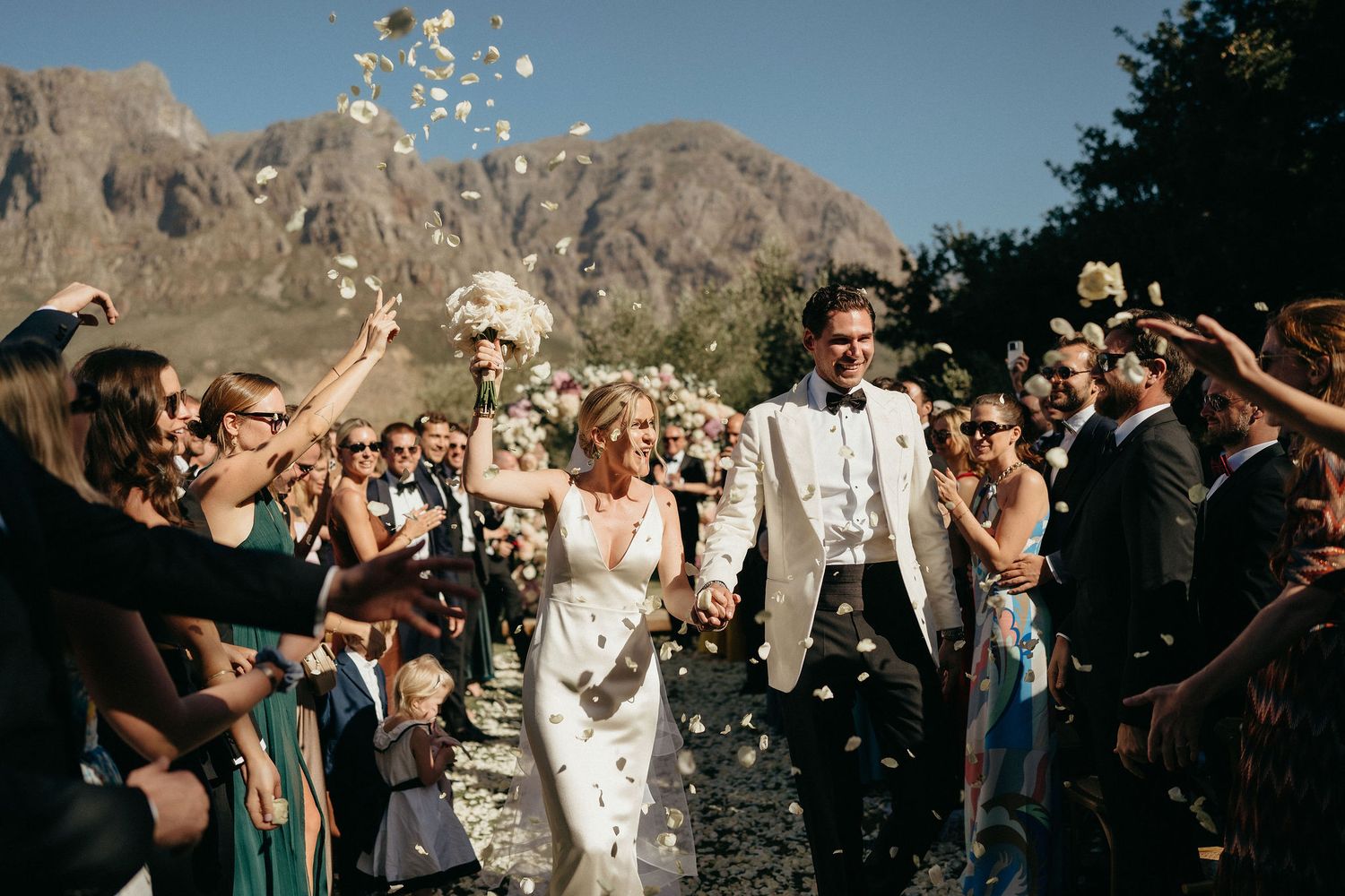 Wedding guests toss confetti in celebration as newlyweds walk through a joyous crowd outdoors.