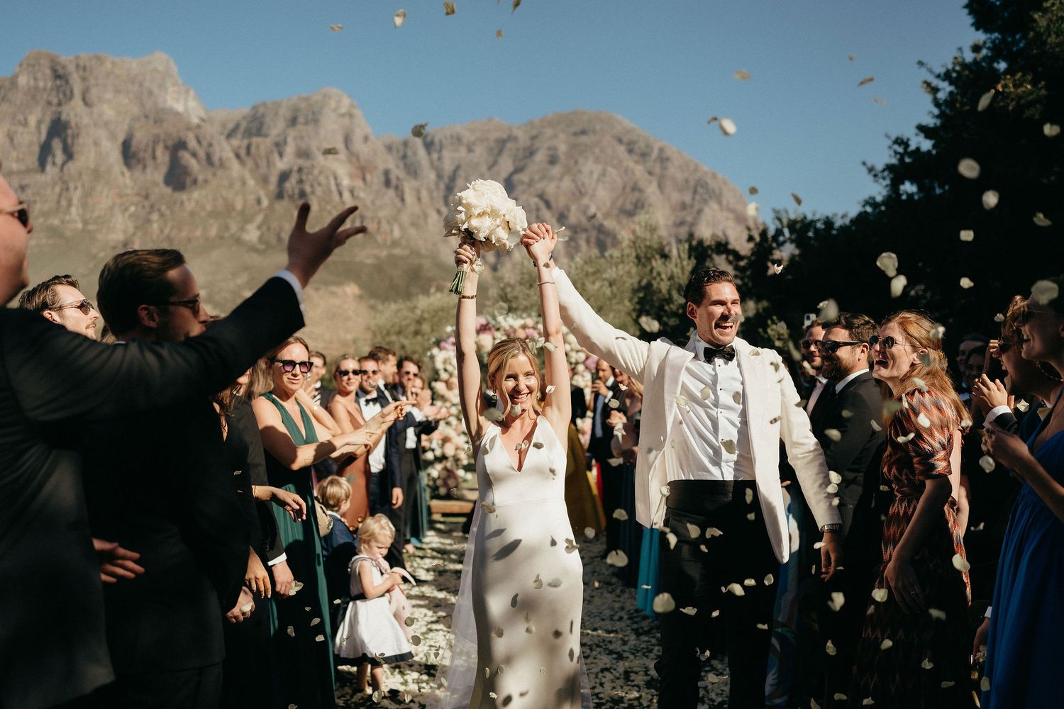 Joyful wedding celebration with guests tossing confetti in front of dramatic mountain backdrop at outdoor ceremony.