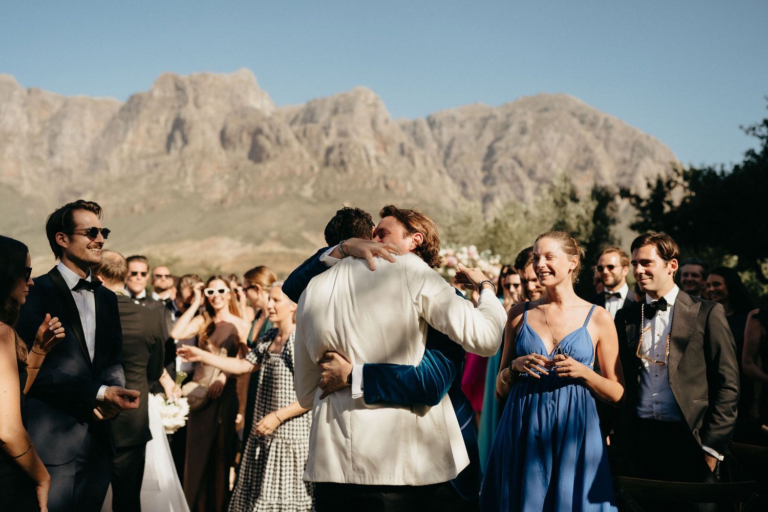 Wedding guests gather outdoors for a celebration with dramatic mountain peaks in the background.