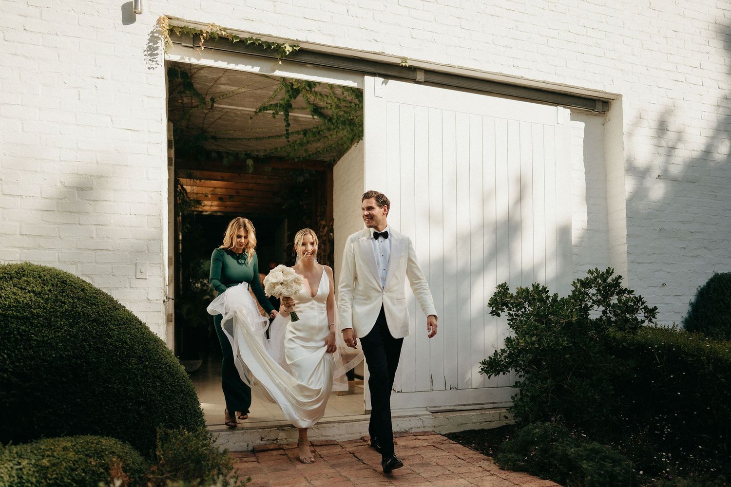 A joyful wedding party exits through white barn doors on a sunny day at an elegant venue.