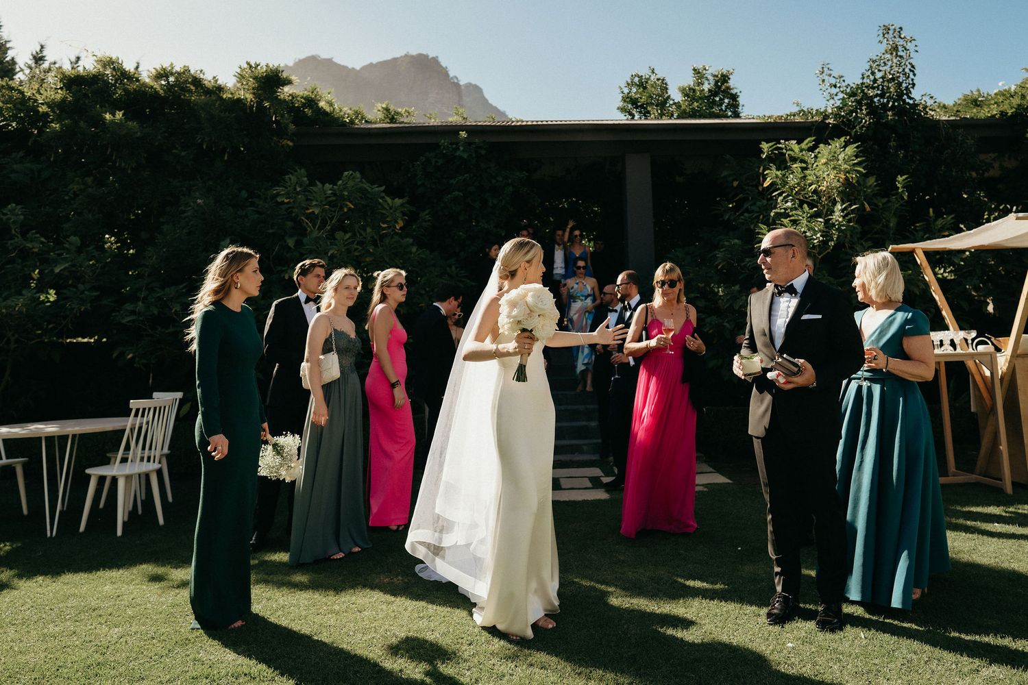 Wedding party members walk in a line across a sunny outdoor lawn during a summer celebration.