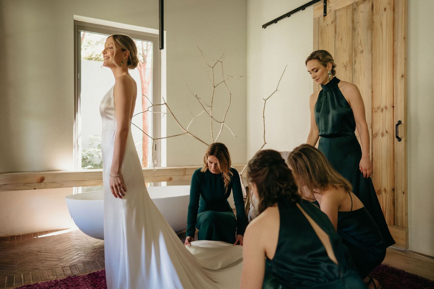 Wedding party gathers in a rustic room as bridesmaids in emerald green dresses help prepare the bride in white.